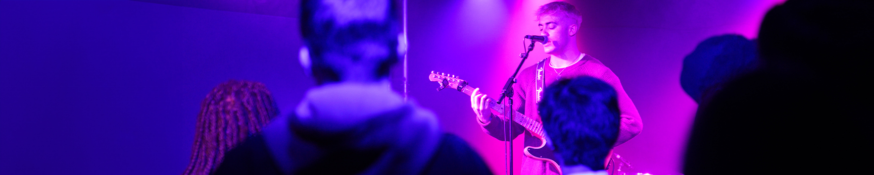 Photograph of a young white male singing into a microphone and playing a guitar with an audience in a room with pink and purple lighting