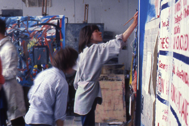 Young student with short dark hair painting in the art school next to an abstract artwork and other students