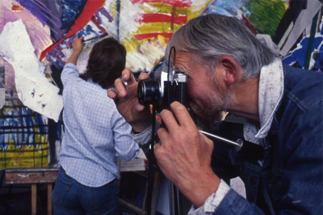 Man with grey hair taking a photo using a black and silver film camera in front of a student working on a large and colourful artwork in the background