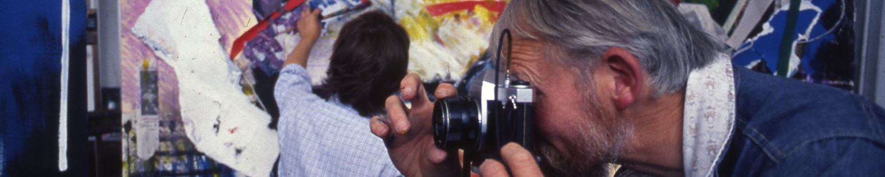 Man with grey hair taking a photo on a black film camera in front of a person painting on a large colourful surface in the background